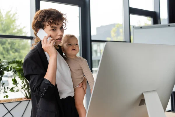 Woman in black suit holding smiling baby while talking on cellphone near computer monitor — Stockfoto