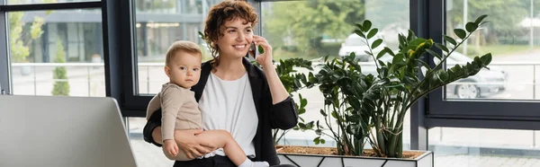 Smiling businesswoman with little daughter talking on smartphone near computer monitor and green plants in office, banner — Photo de stock