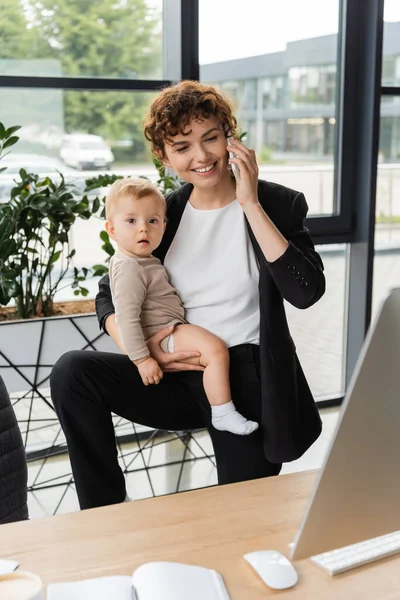 Joyful businesswoman holding toddler daughter and talking on smartphone near computer monitor in office — Stockfoto