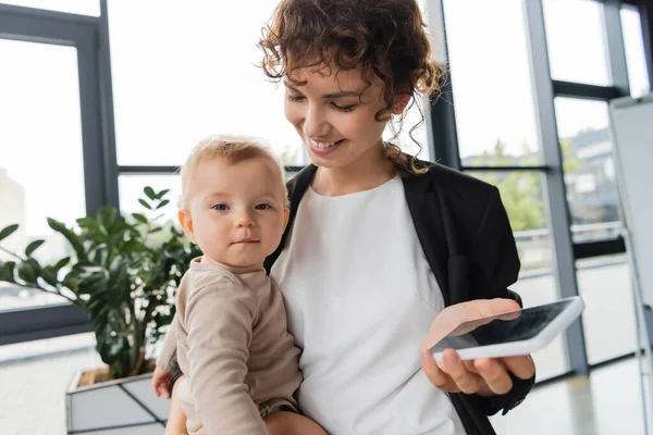 Little baby looking at camera near smiling mother with mobile phone in office — Stockfoto