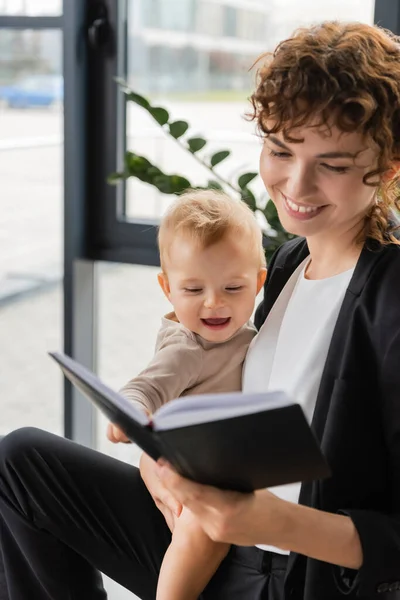 Smiling businesswoman holding cheerful toddler daughter and looking in notebook in office - foto de stock
