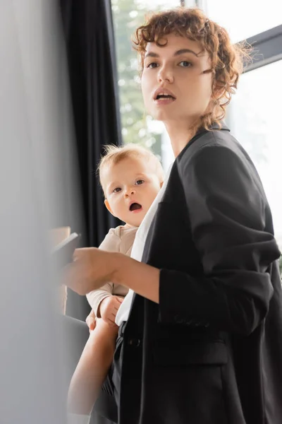 Woman in black blazer looking away while standing with toddler daughter in office on blurred foreground — Photo de stock