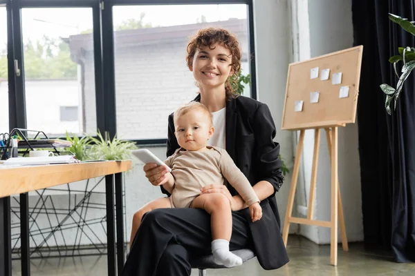 Happy businesswoman sitting with little daughter and mobile phone while looking at camera in office — Photo de stock