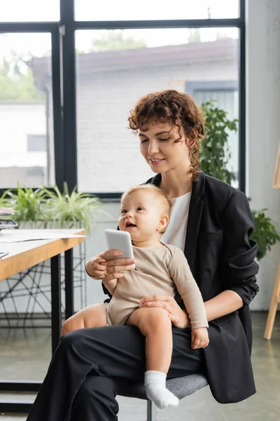 Smiling businesswoman in black suit using mobile phone while sitting with toddler daughter in office — Stock Photo