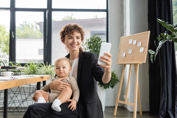 Joyful businesswoman in black suit sitting with baby and taking selfie on cellphone in office — Foto stock