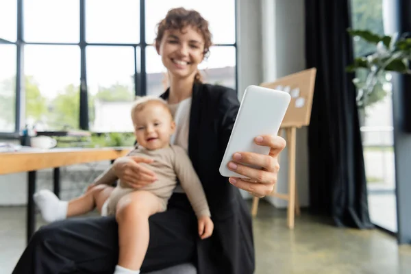 Blurred businesswoman taking selfie on mobile phone while sitting with smiling baby in office — Stock Photo