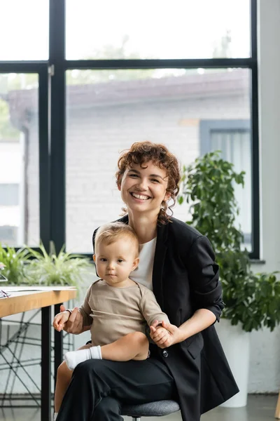 Щаслива бізнес-леді, сидячи з дитиною в сучасному офісі і дивлячись на камеру — стокове фото