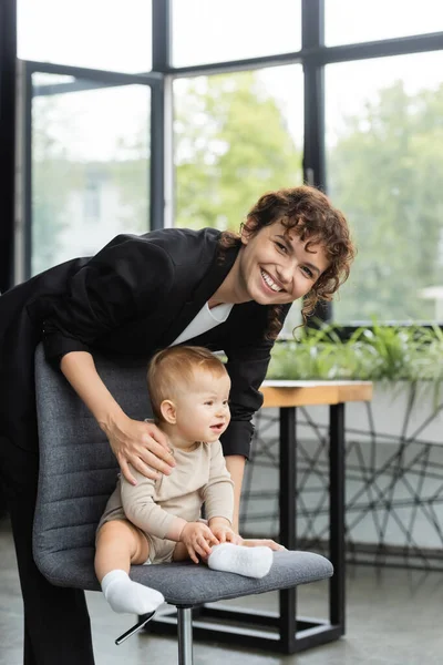 Cheerful businesswoman smiling at camera near toddler child sitting on office chair — Photo de stock