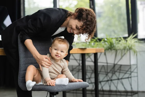 Весёлый ребёнок в комбинезоне сидит на офисном стуле рядом с улыбающейся матерью в официальной одежде — стоковое фото