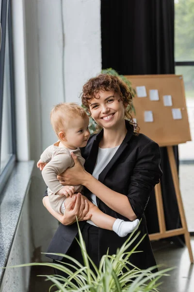 Joyful businesswoman in black blazer looking at camera while standing with toddler daughter in office - foto de stock