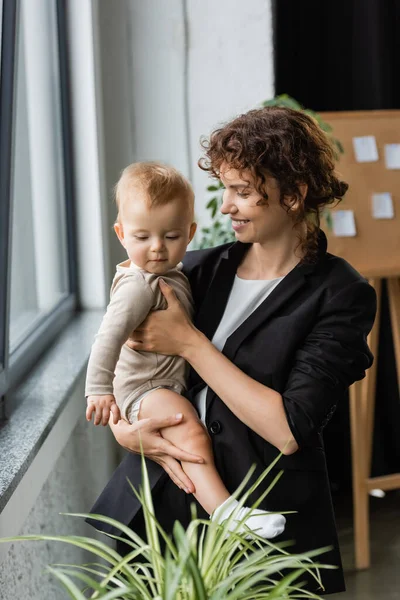 Smiling businesswoman in black blazer standing with toddler child near blurred plant in office - foto de stock