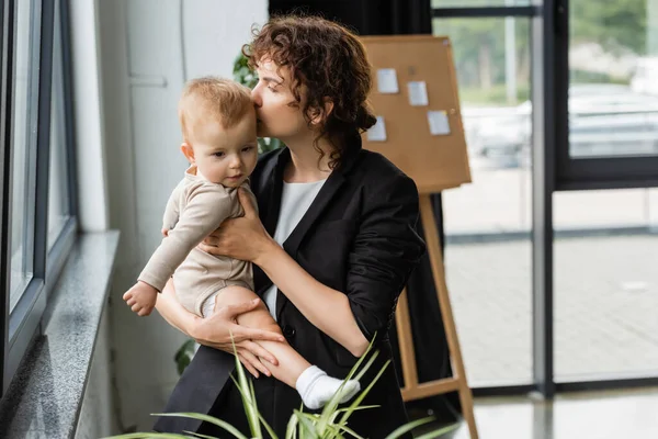 Бізнес-леді в чорному блістері цілує доньку малюка в офісі — стокове фото