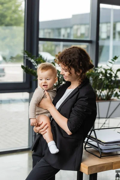 Businesswoman in black suit holding little child near work desk with documents in office — Stock Photo