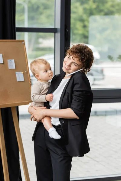 Positive businesswoman in black suit holding little daughter and talking on smartphone in office — Stock Photo
