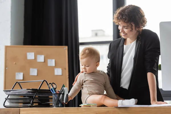 Businesswoman smiling near baby sitting on office desk near cork board with sticky notes on blurred background - foto de stock