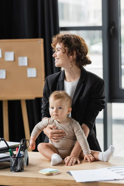 Positive woman in black formal wear looking away near little daughter sitting on office desk near documents — Stock Photo