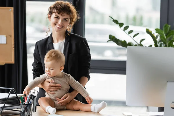 Businesswoman in black blazer smiling at camera near little daughter in romper sitting on office desk — Photo de stock