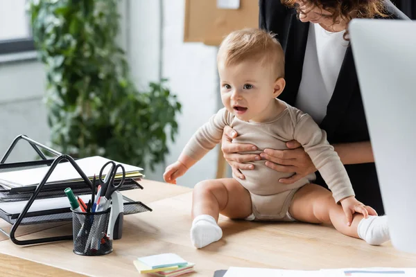 Businesswoman holding excited baby sitting on office desk near documents and stationery — Photo de stock