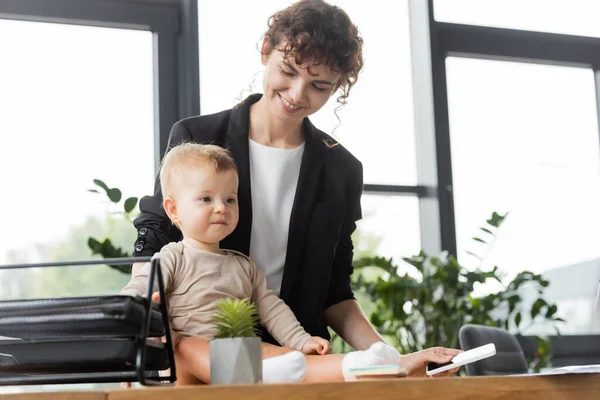 Happy businesswoman in black blazer holding mobile phone near toddler daughter sitting on office desk — Photo de stock