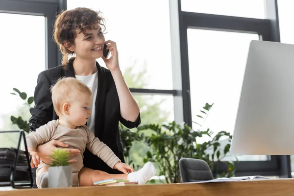 Toddler girl sitting on office desk near happy mother talking on smartphone in office — Photo de stock