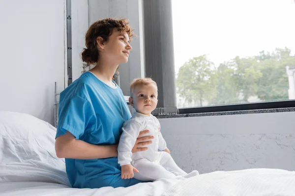 Smiling woman in patient gown sitting with toddler daughter on hospital bed near window — Stock Photo