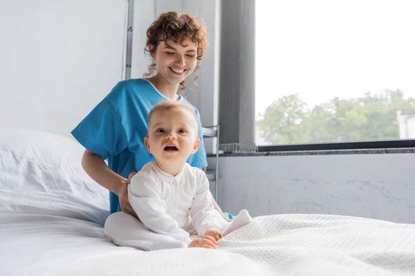 Toddler child with open mouth sitting on hospital bed near smiling mom in patient gown — Stock Photo