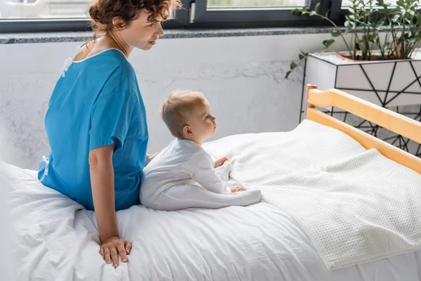 Side view of woman in patient gown and toddler girl in romper sitting on bed in clinic — Stock Photo