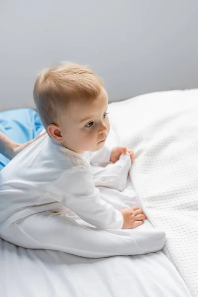 Little baby in white romper looking away while sitting near mom on bed in hospital ward — Stock Photo