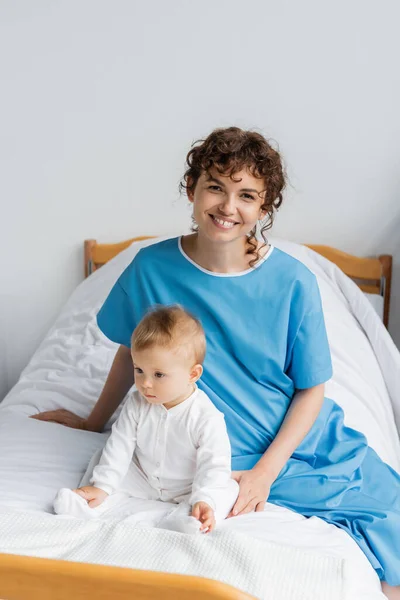 Cheerful woman in hospital gown looking at camera while sitting near baby on hospital bed — Stock Photo