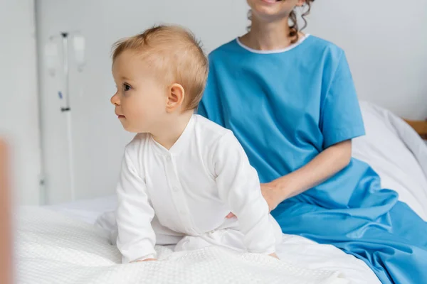 Baby girl in romper looking away while crawling on hospital bed near mom in patient gown — Stock Photo