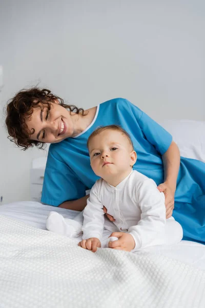 Toddler child sitting on hospital bed and looking at camera near smiling mother in patient gown — Stock Photo