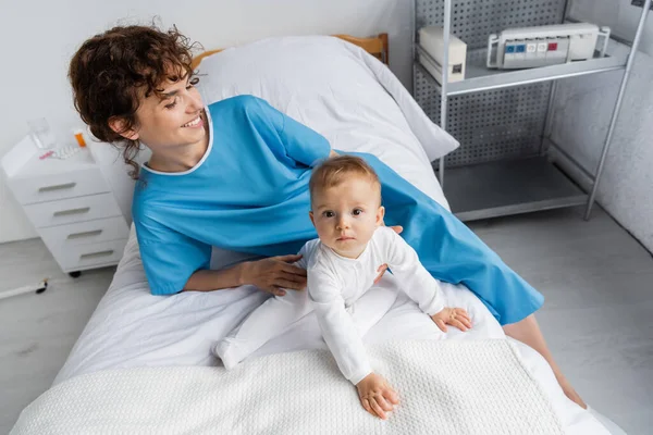 High angle view of baby in romper looking at camera near smiling mom on hospital bed — Stock Photo