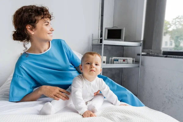 Toddler child in romper sitting near mom in patient gown looking away on bed in clinic — Stock Photo