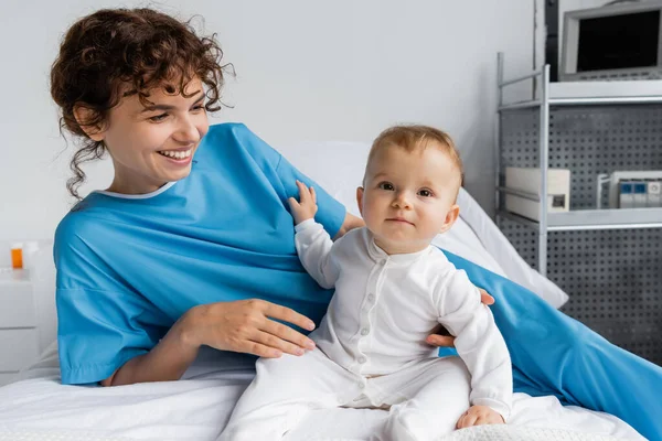 Joyful woman in patient gown looking at baby sitting in romper on hospital bed — Stock Photo
