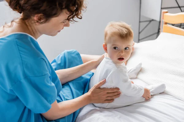 Baby girl with open mouth looking away while sitting near mom on bed in clinic — Stock Photo