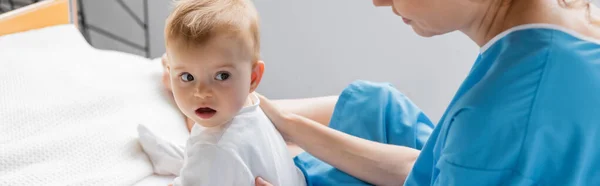 Toddler girl looking away while sitting with open mouth near mother on hospital bed, banner — Stock Photo