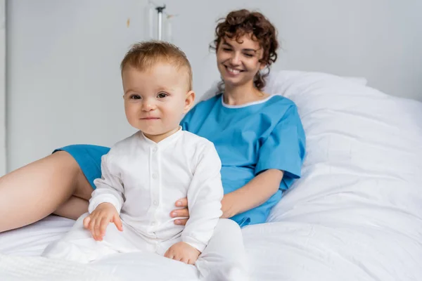 Baby girl in romper looking at camera while sitting on hospital bed near mom smiling on blurred background — Stock Photo