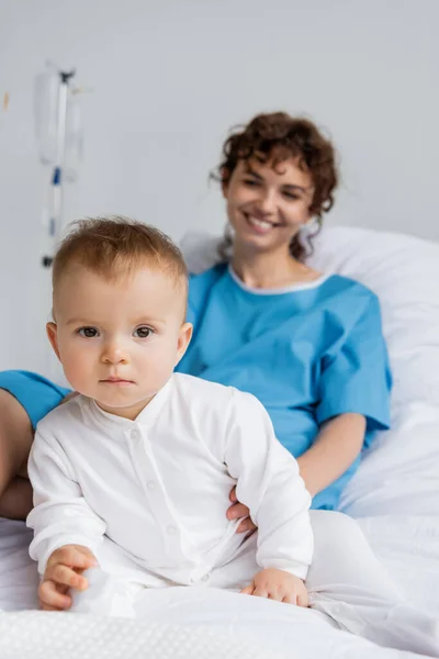 Toddler girl in romper looking at camera near blurred mother smiling on hospital bed — Stock Photo