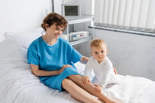Woman in patient gown smiling near little daughter on bed in hospital ward — Stock Photo