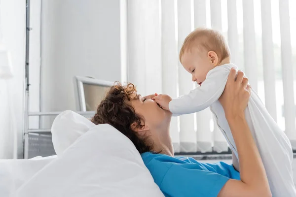 Side view of little girl touching face of happy mother lying on hospital bed — Stock Photo