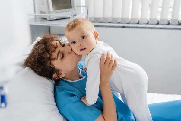 Woman with closed eyes embracing and kissing little daughter in romper on bed in hospital ward - foto de stock