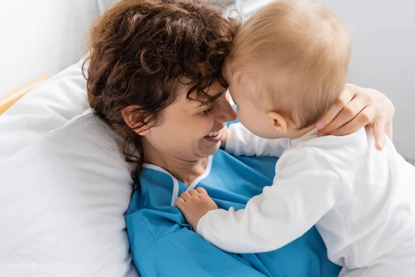 Cheerful woman embracing toddler daughter while lying on bed in clinic - foto de stock