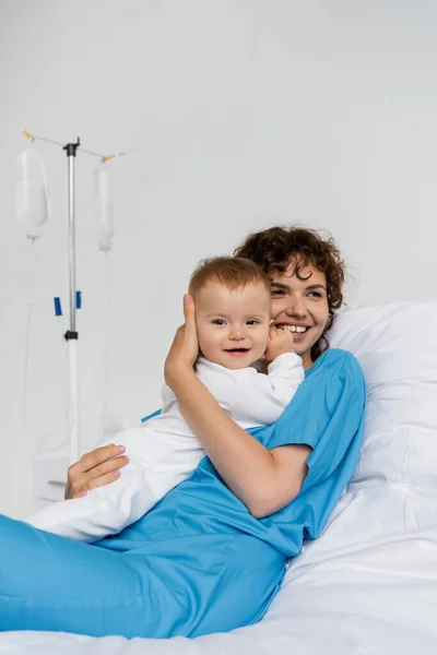 Joyful woman embracing baby in romper while looking away on bed in clinic — Stock Photo