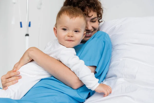 Happy woman in patient gown embracing little daughter on bed in clinic — Stock Photo