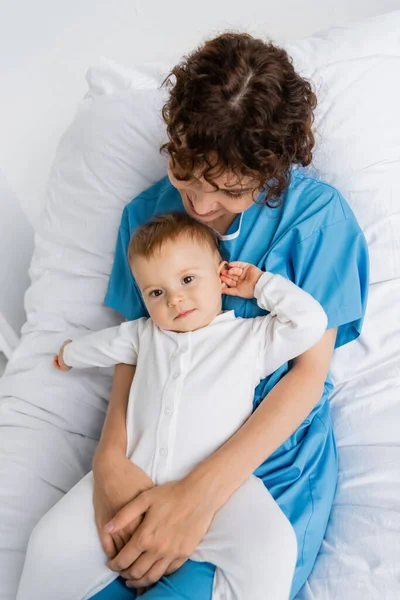 Top view of woman in patient gown holding toddler daughter in romper on hospital bed — Stock Photo