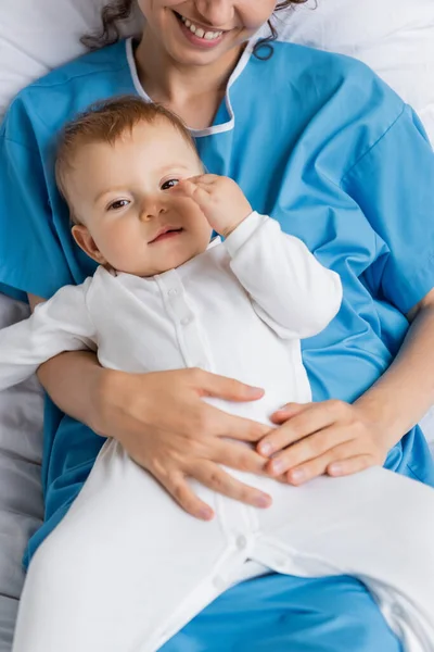 Top view of baby in romper looking at camera near smiling mother in patient gown — Stock Photo