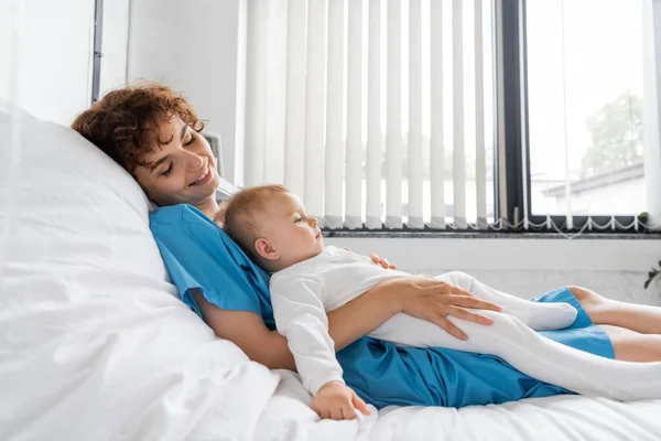 Pleased woman in patient gown lying on hospital bed with toddler child in romper — Stock Photo