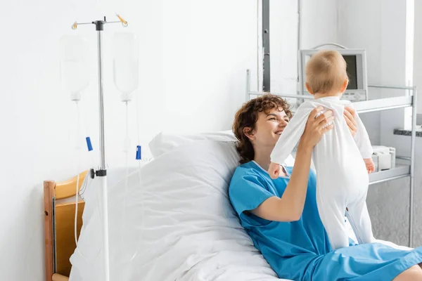 Happy woman in patient gown holding baby in romper while lying on bed in hospital ward — Stock Photo