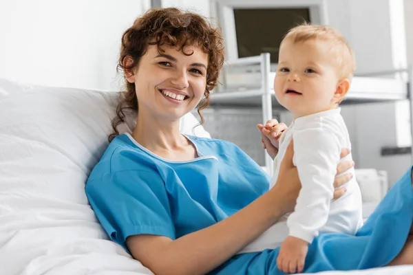 Joyful woman in patient gown looking at camera near little daughter in hospital ward — Stock Photo
