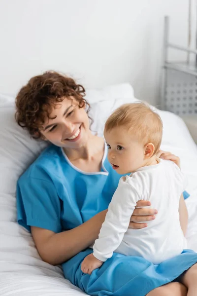 Joyful woman in patient gown lying on hospital bed and holding toddler daughter in romper — Stock Photo